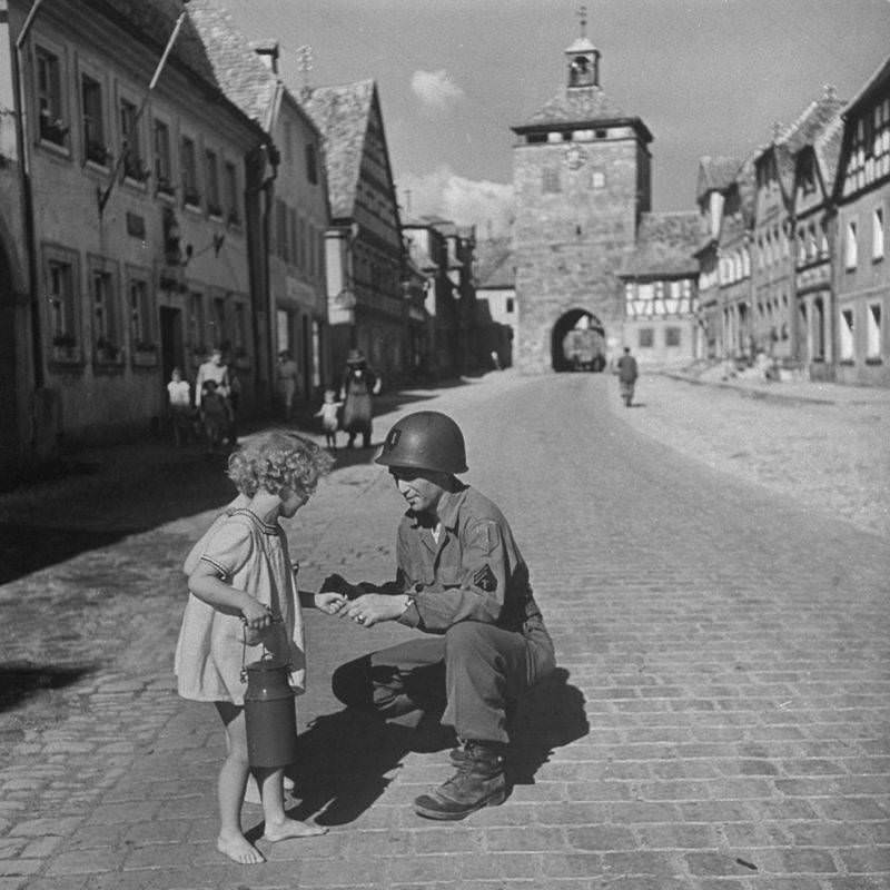 #56 American Supply Sergeant Ralph Gordon kneels in a street to give a piece of gum to a barefoot German girl, 1945.