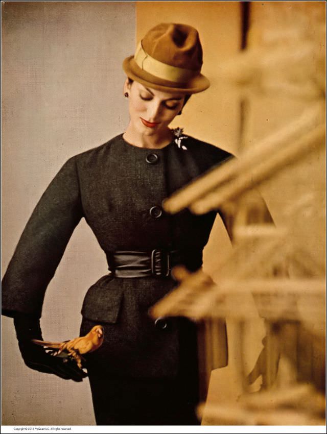 #63 Carmen in smoky charcoal suit of British wool tweed, tunic jacket is collarless and belted in black kidskin, by Ben Zuckerman, hat by Emme, 1959.