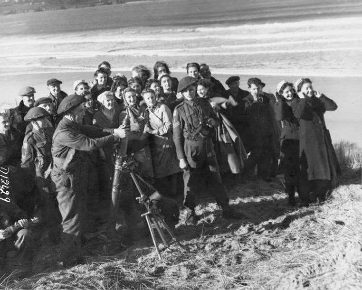 #1 A group of mostly women war workers stand, many with their fingers in their ears, on the sand of a coastal test range to watch as several soldiers launch mortar bombs, 1943.