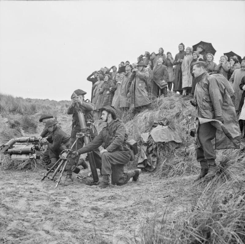 #59 A group of war workers stand, on a grassy mound at a coastal test range to watch as soldiers launch mortar bombs from the beach immediately below them, 1943.