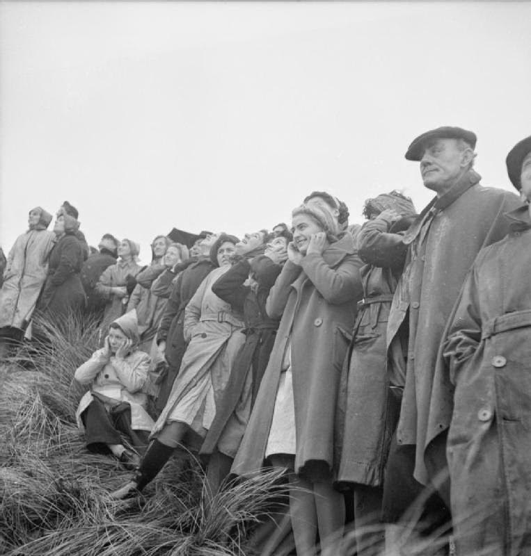 #2 A group of war workers stand in a row on a grassy hill on a coastal test range to watch mortar bombs being launched, 1943.