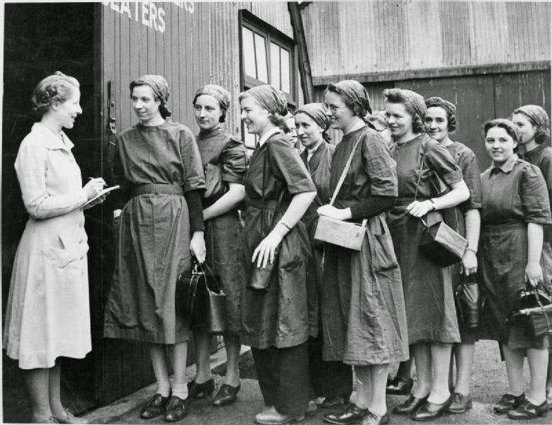 #30 A group of new recruits dressed in overalls and head scarves and carrying their gas masks are met by the female supervisor at Slough Training Centre as they enter the workshop for the first time, 1941.
