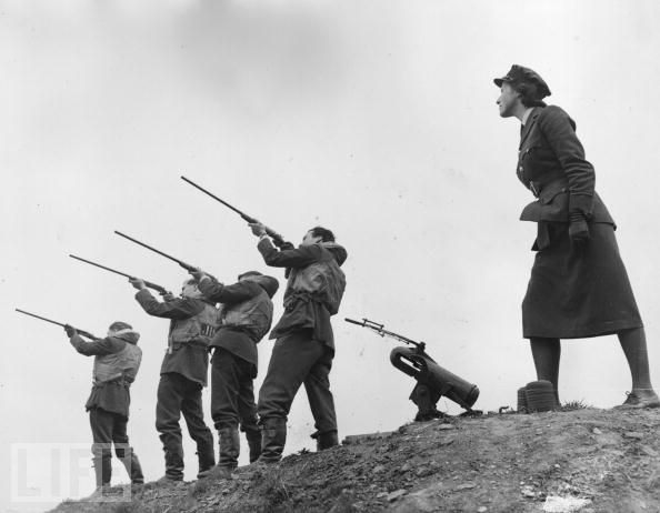 #48 Pilots of a Spitfire fighter squadron, wearing their life-vests to be ready to take off at any time, take part in a clay pigeon shoot. An officer of the Women’s Auxiliary Air Force assists them by operating the clay pigeon trap.