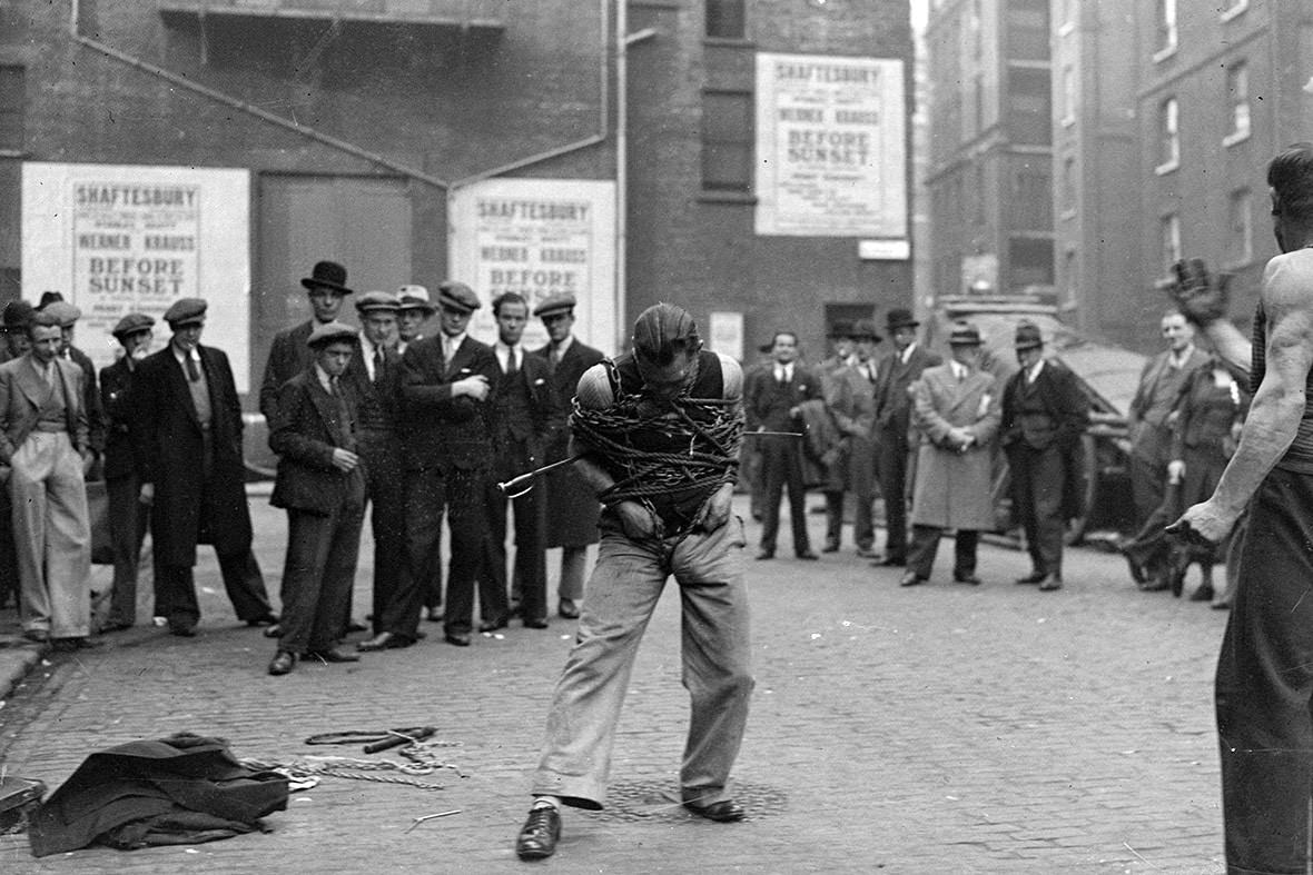#7 A small crowd gathers to watch a man trying to extract himself from chains on a street in Soho, 1937