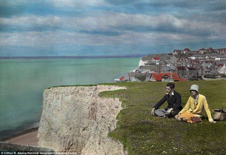 #62 Two young adults sit in the grass by a cliff near water and the village of Rottingdean in East Sussex in 1931