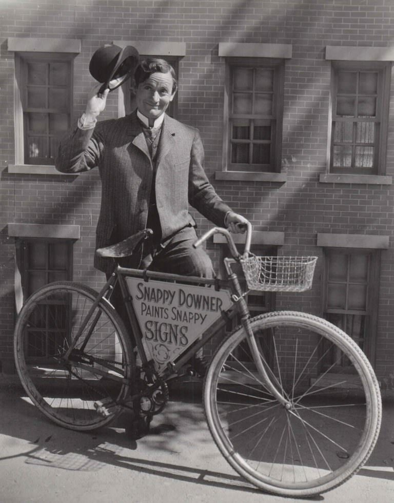 #14 Roscoe Karns posing with a bike, tips his hat.