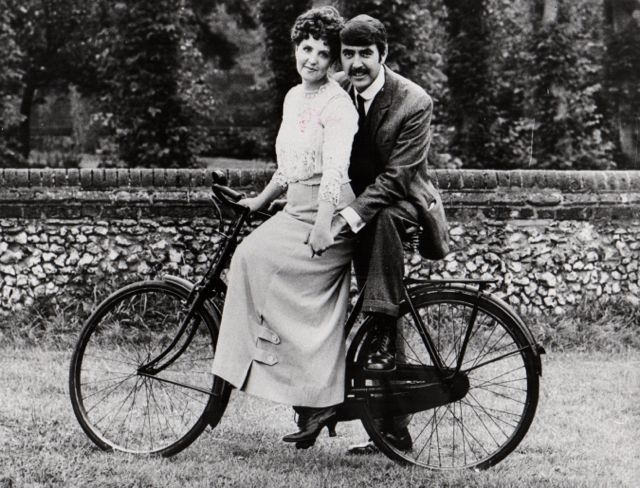 #41 Pauline Collins and John Alderton posing on a bike.