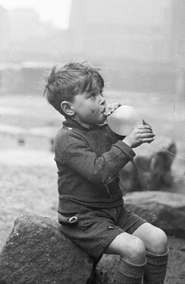 #37 A young boy sits on a stone and attempts to blow up his balloon in the Gorbals