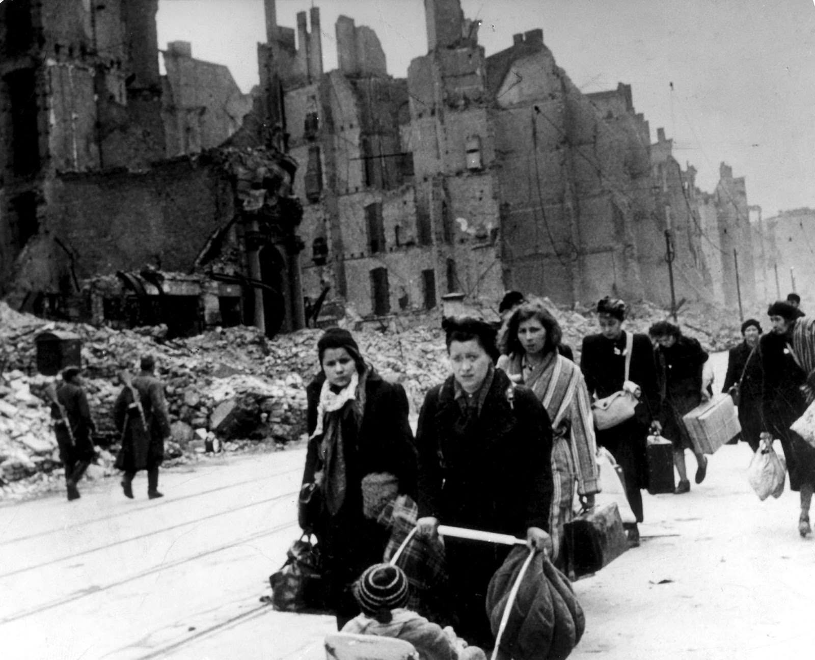 #21 A stream of refugees and people who have been bombed out of their homes moving through destroyed streets in Germany in 1945, after end of war. On the left, two Soviet soldiers can be seen patrolling.