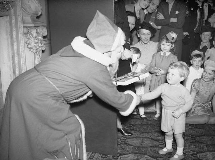 #73 Admiralty Christmas Party for Allied Naval Officers’ children, 1942. This photograph shows ‘Father Christmas’ presenting Winston Churchill Junior, the Prime Minister’s grandson, with a gift of a book of nursery rhymes at a Christmas party at Admiralty House.