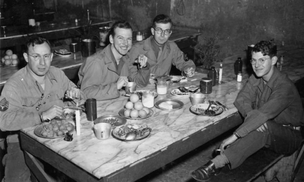 #68 British soldier eating Christmas meal in his slit trench w. holiday cards from home propped up outside, Netherlands, 1944.