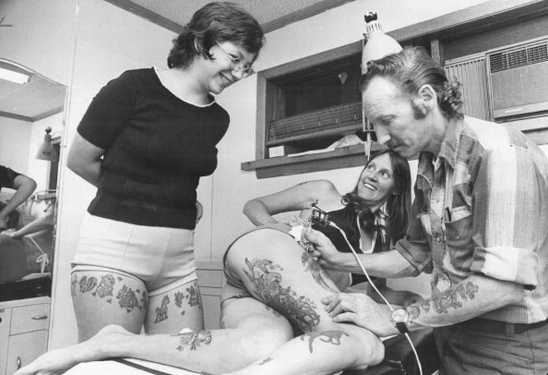 #18 Beachcomber Bill’s wife; Brenda; smiles from table as he shows how he applies tattoo, 1975.