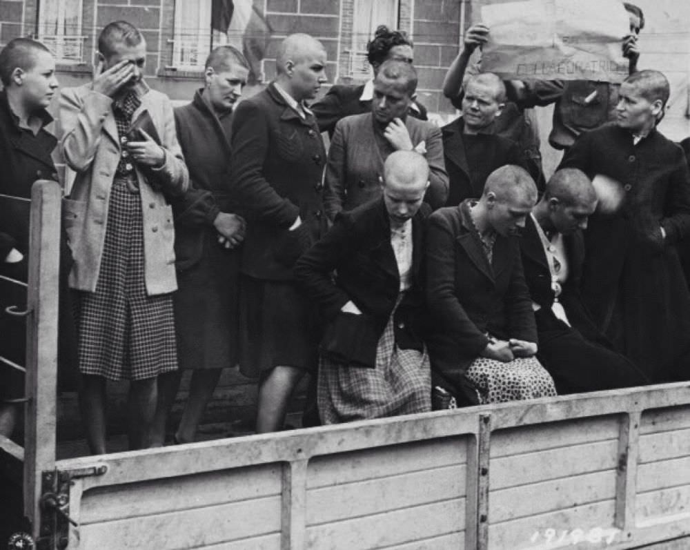 Brutalized women, their heads shaved, are loaded into the back of a truck. The ‘man’ behind them is holding a sign that says ‘collaborators’, but who is the real collaborator?