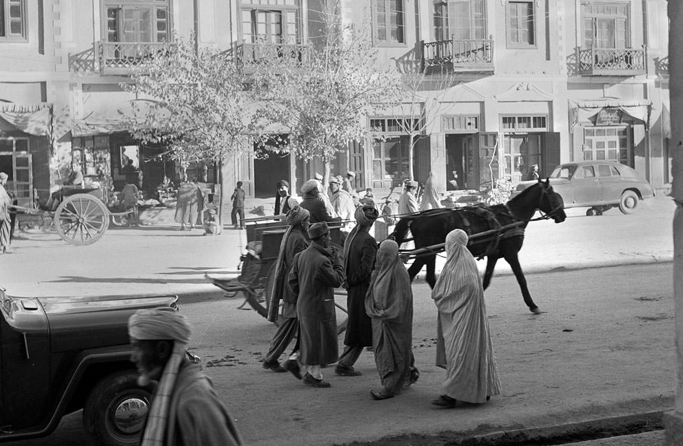 #27 Women, wearing traditional burqas and Persian slippers, walk alongside men, cars and horse carts, in a street in Kabul, in 1951. At the time, this street was one of only three paved streets in the capital city.