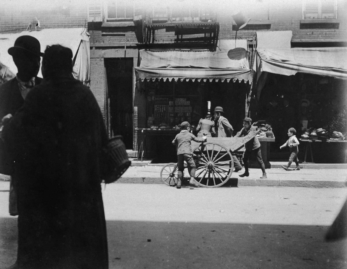 #20 Two young boys laugh and steal items from a vendor’s pushcart on Hester Street in the Lower East Side, 1895