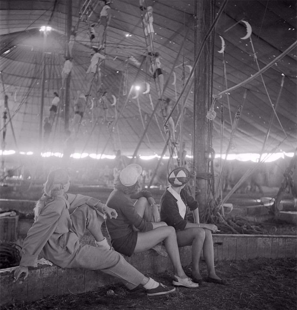 #15 Three circus girls sitting on a wooden plank and watching a group of aerialists rehearsing with ropes for the Ringling Bros. and Barnum & Bailey Circus in Sarasota, FL in 1949.