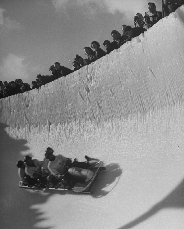 #7 Good of cresta, a kind of bobsled, coasting around sunny bend as people peer from above the cresta run.