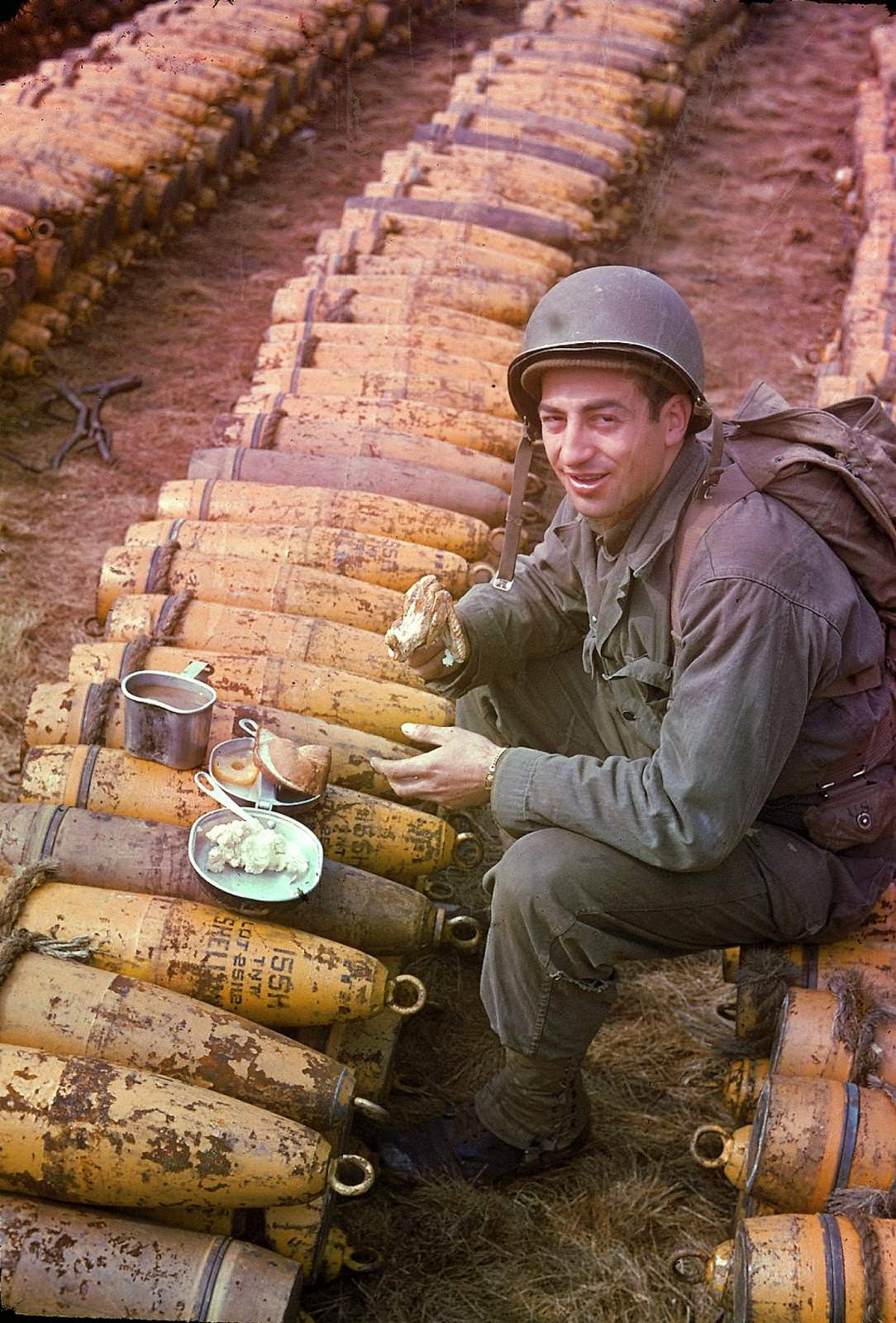 #11 American soldier sits and eats his meal (which includes chicken, mashed potatoes, bread, and pineapple) atop rows of a stockpiled ammunition shells, England, May 1944.