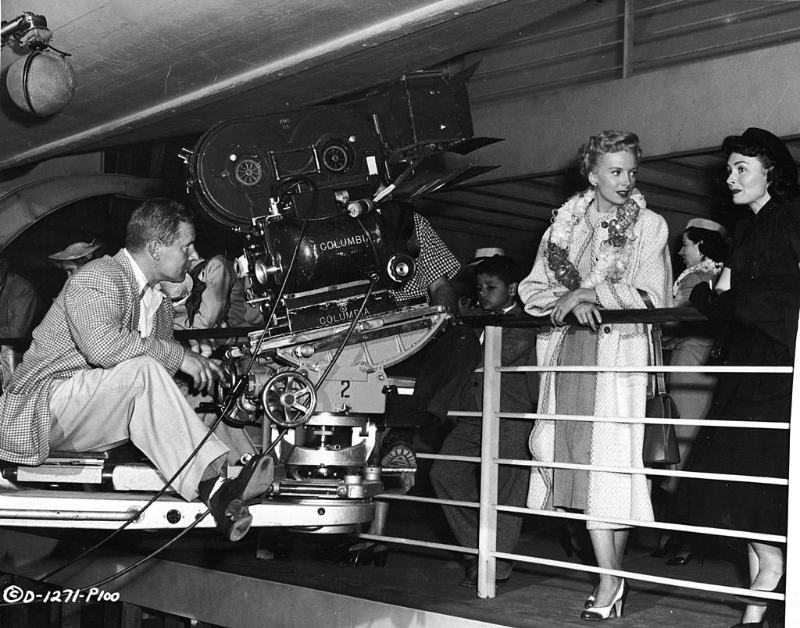 Deborah Kerr and Donna Reed standing by a railing while cameraman Kit Carson rolls film on the set of ‘From Here To Eternity,’ 1953