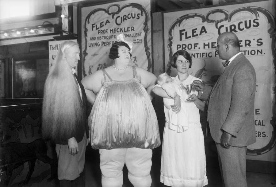 At a Coney Island “freak” show an albino is photographed with the Fat Lady. A Flea Circus poster is in the background.