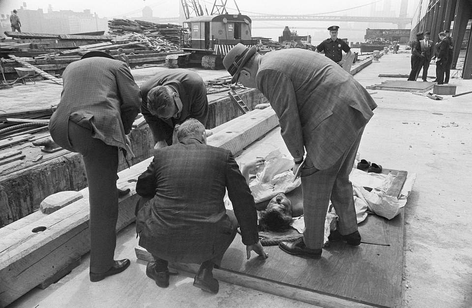 A medical examiner peering over the body of a man found floating in the East River in 1963. Bullet holes were found in the man’s head