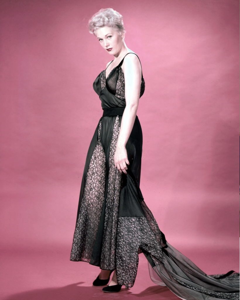 #8 Kim Novak wearing a long black silk and black lace dress, with a black lace train, in a studio portrait, against a red background, circa 1960