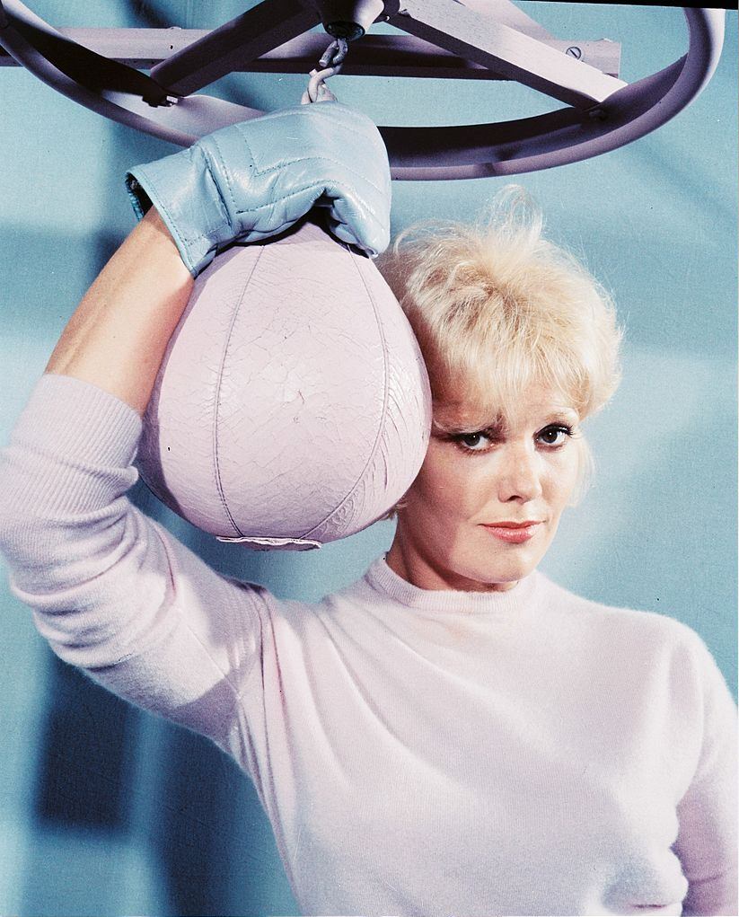 #57 Kim Novak wearing a white woollen jumper and blue boxing gloves poses with a white punch ball, circa 1965