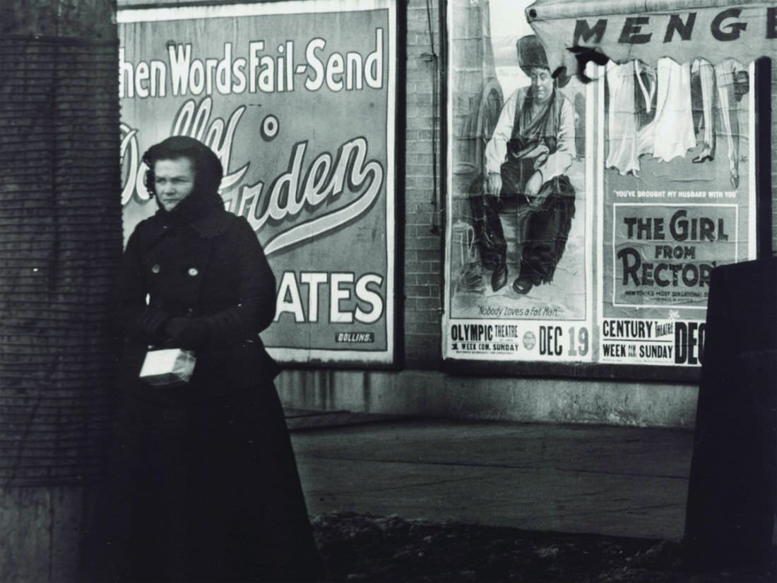 #69 A woman bundled in winter wear at an unidentified street corner in front of advertisements for the movies The Round Up at the Olympic Theatre and The Girl from Rector’s showing at the Century Theatre, 1917
