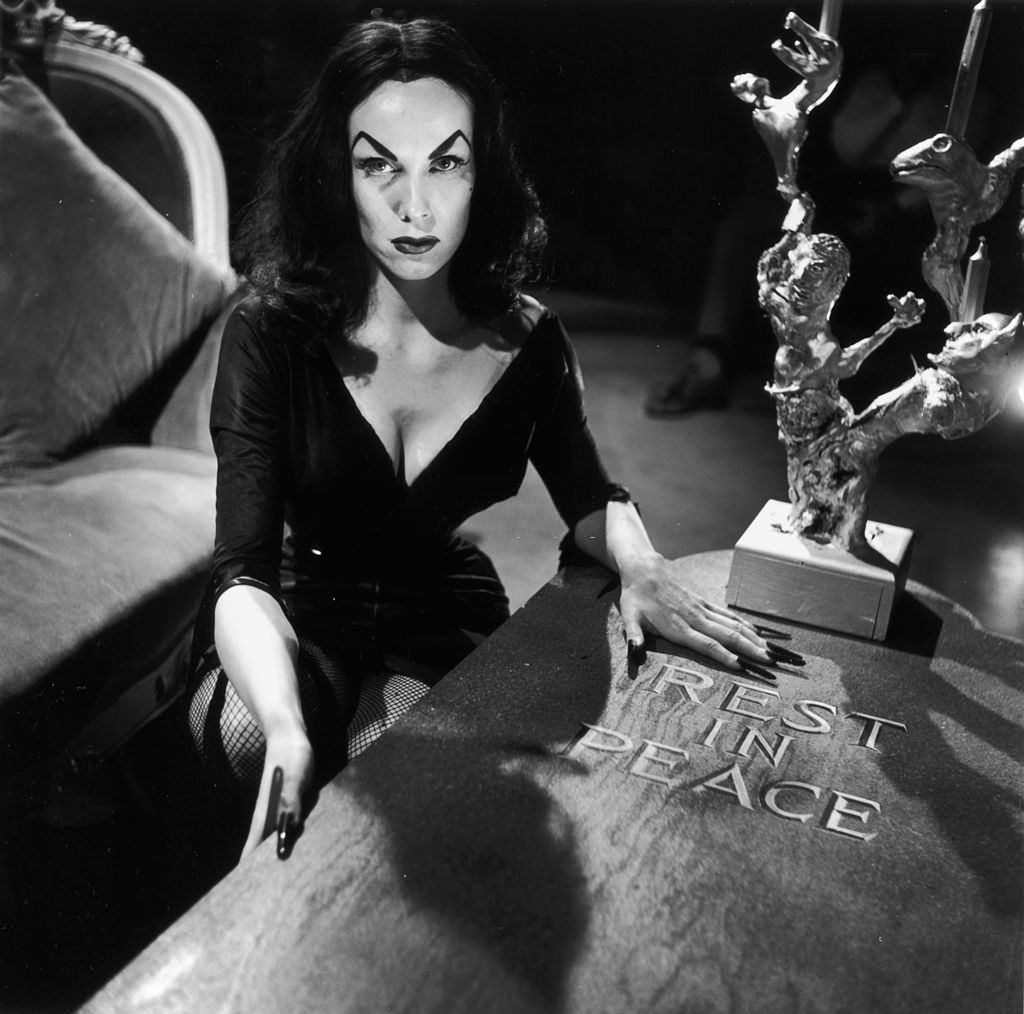 Maila Nurmi crouching beside a wooden coffin marked ‘Rest in Peace’. She has arched, painted eyebrows and wears a low-cut black dress with fishnet stockings, 1956