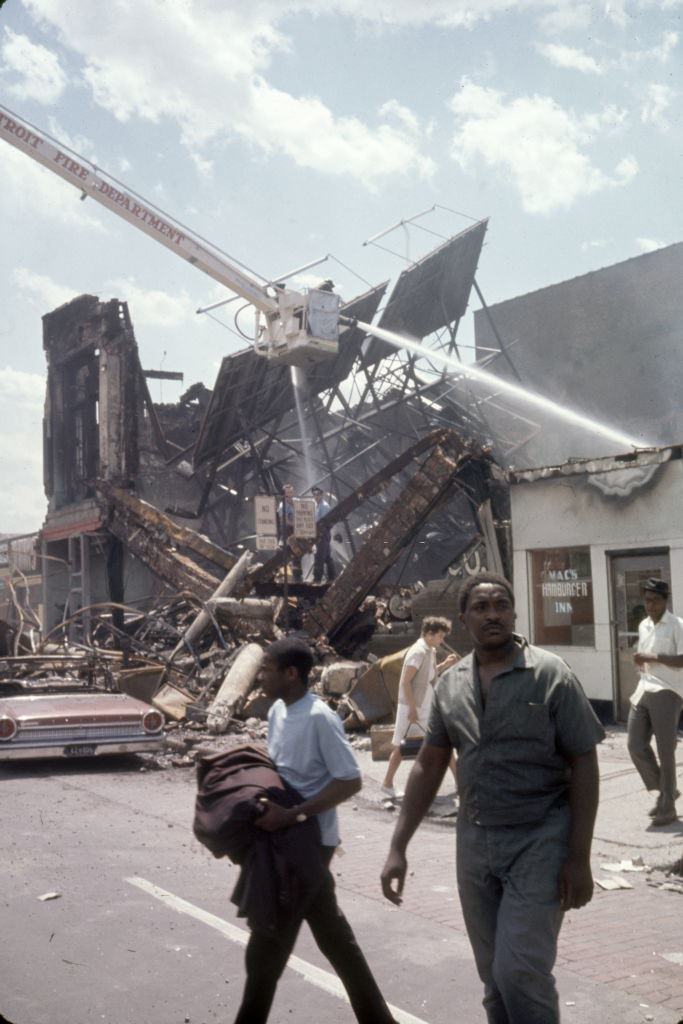 #11 View of a firefighters as they battle a blaze as pedestrians walk beneath them in the wake of ongoing riots, Detroit, Michigan, 1967