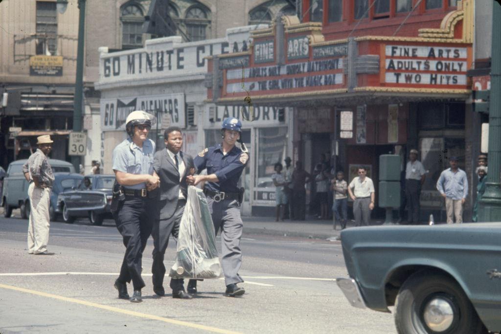 #12 A pair of police officers arrest a young man, dressed in a suit, and escort him down the middle of Woodward Avenue, Detroit, 1967