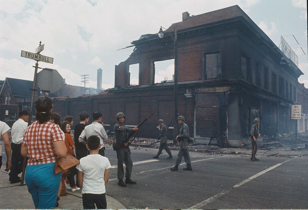 #27 Bystanders observe National Guardsmen, armed with rifles standing guard and patrolling a street corner beside a burnt out brick building on the West Side of Detroit, 1967