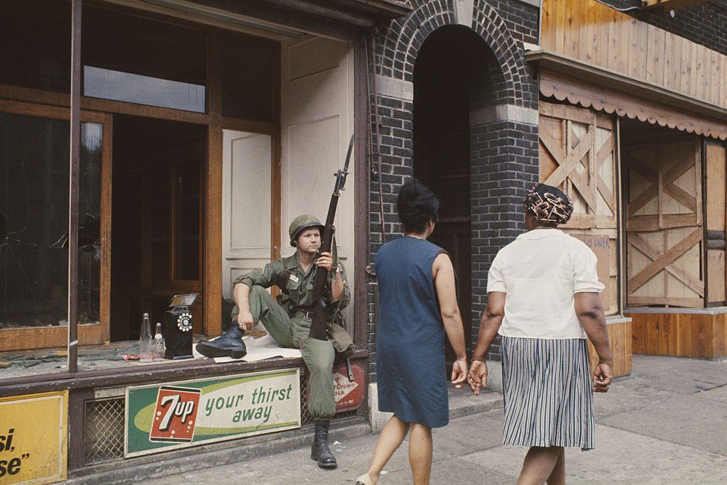 #28 A National Guardsman armed with a rifle and bayonet rests in the window of a wrecked shop front in a shopping street in Detroit, 1967