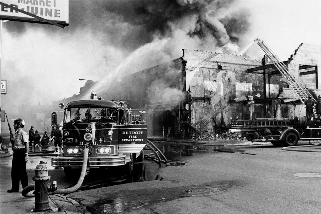 #39 A policeman stands guard in a Detroit street on July 25, 1967 as firefighters try to extinguish a burning building during riots that erupted in Detroit