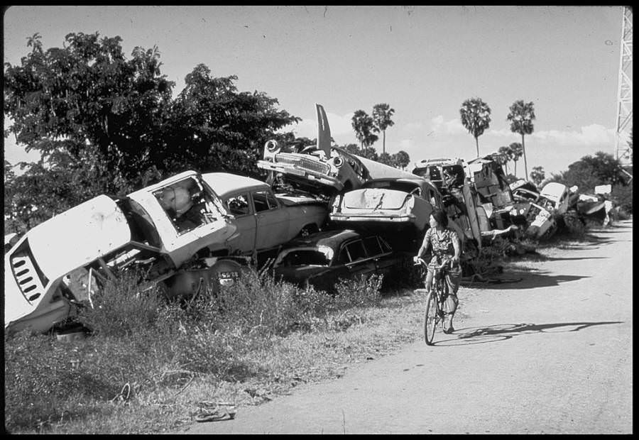#22 A woman rides a bicycle by a stack of destroyed cars, cast aside by the Khmer Rouge as of symbol of the bourgeoisie, 1979