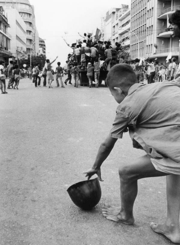 #36 A young boy picks up a soldier’s helmet as the victorious Khmer Rouge parades through the streets of his city, Phnom Penh, 1975.