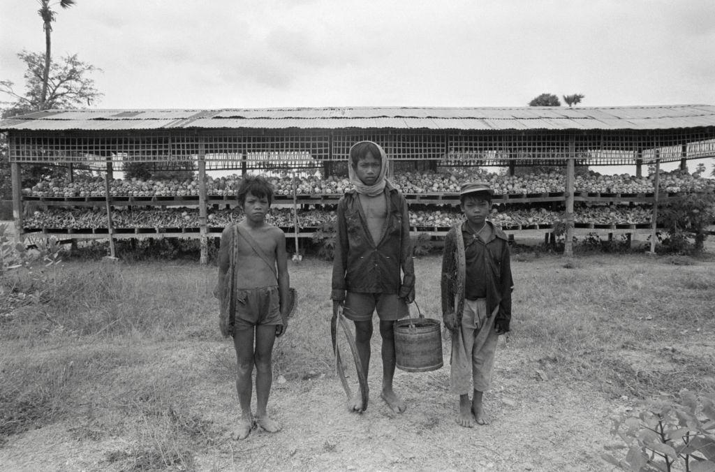 #8 Mass Grave in Cambodia. The simple hut behind the three boys at the Choeung Ek extermination camp contains hundreds of human skulls and bones.