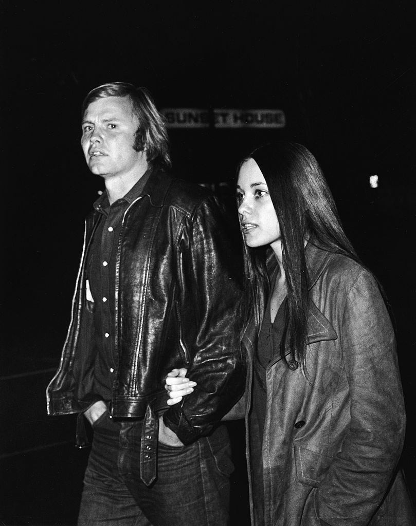 #10 Marcheline Bertrand with Jon Voight walk hand-on-arm along a street at night in front of a sign which reads ‘Sunset House,’ 1970s.