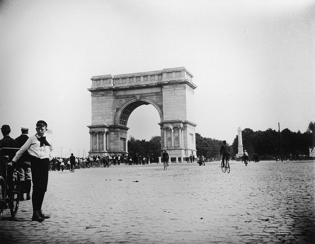#33 A boy leans against a bicycle in the foreground as people in the background ride their bikes through the arch at Prospect Park during a bicycle parade, Brooklyn, New York, June 15, 1895.