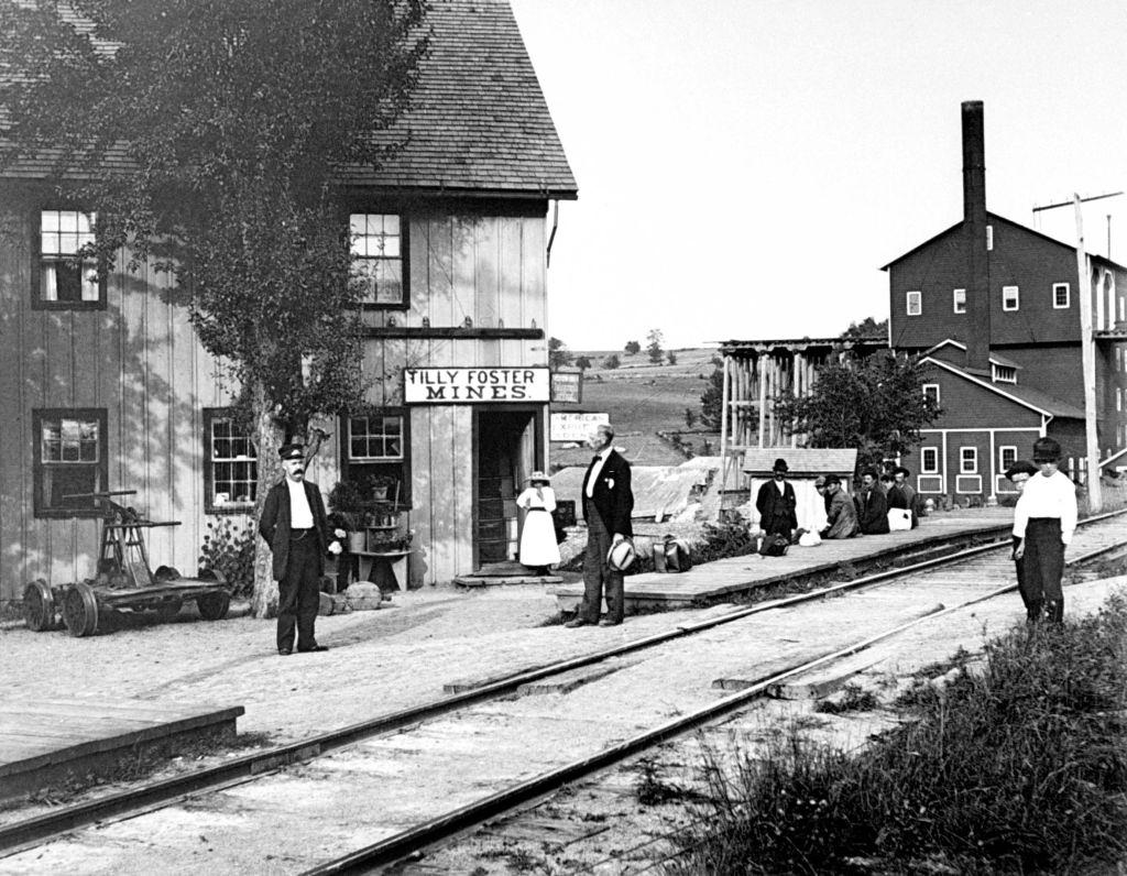 #34 People hanging around outside railroad station, which has signs on it saying TILLY FOSTER MINES, New York, 1889