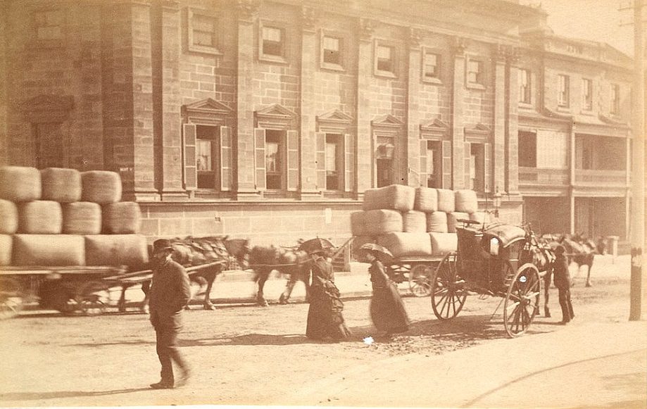 #15 Horses and carts stacked with what appear to be bales of wool being transported through the city, while women in long dresses shaded with umbrellas cross the street, circa 1885-1890.