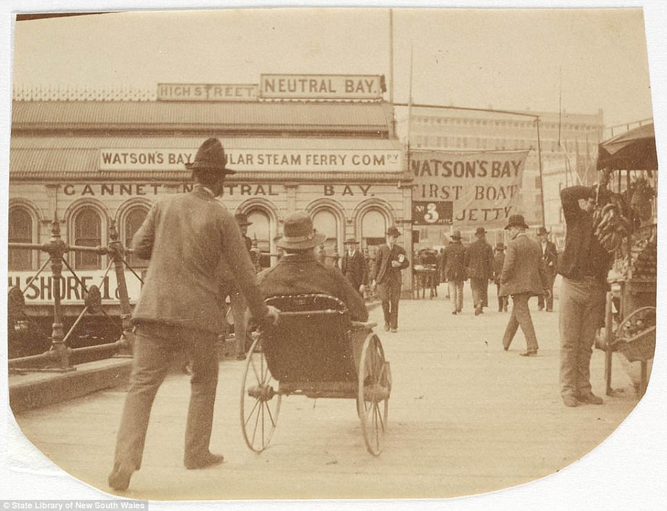 #1 A men pushing another person along in a wheelchair at Circular Quay, circa 1885-1890. Signs advertising the ferry to Watsons Bay are visible, as vendors sell bananas and fruit from stalls on the side of the street .