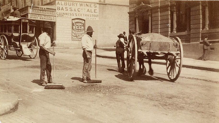#34 Two men sweeping the dirt street, while horse and carriages and horse and carts trundle past, circa 1885-1890.