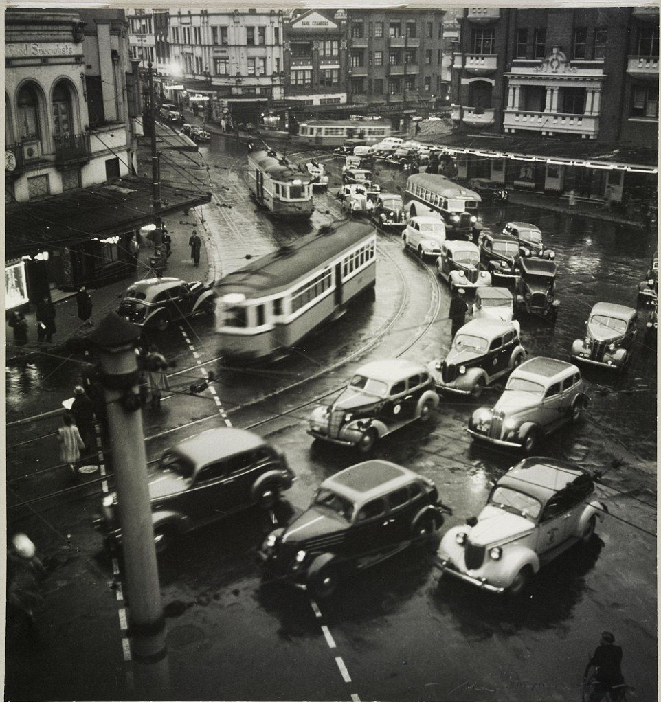 Trams and automobiles racing through the streets and around a corner during rush hour in Kings Cross in 1938