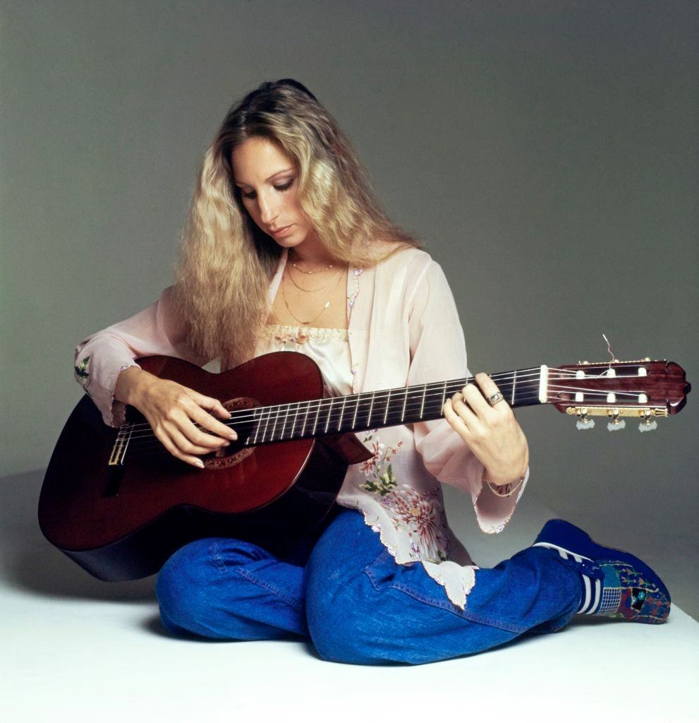 #58 Barbra Streisand wearing blue jeans and a Balinese jacket and playing a guitar, 1975
