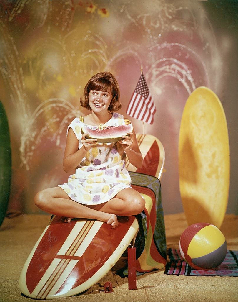 #30 Sally Field sitting on a wooden surfboard with an American flag and eating a watermelon, for the television series, ‘Gidget,’ 1965