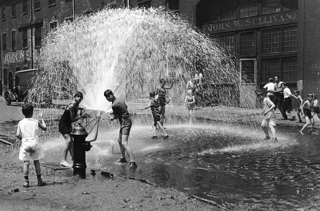 #43 A group of children in shorts play in an open fire hydrant on a cobblestone street, New York City.