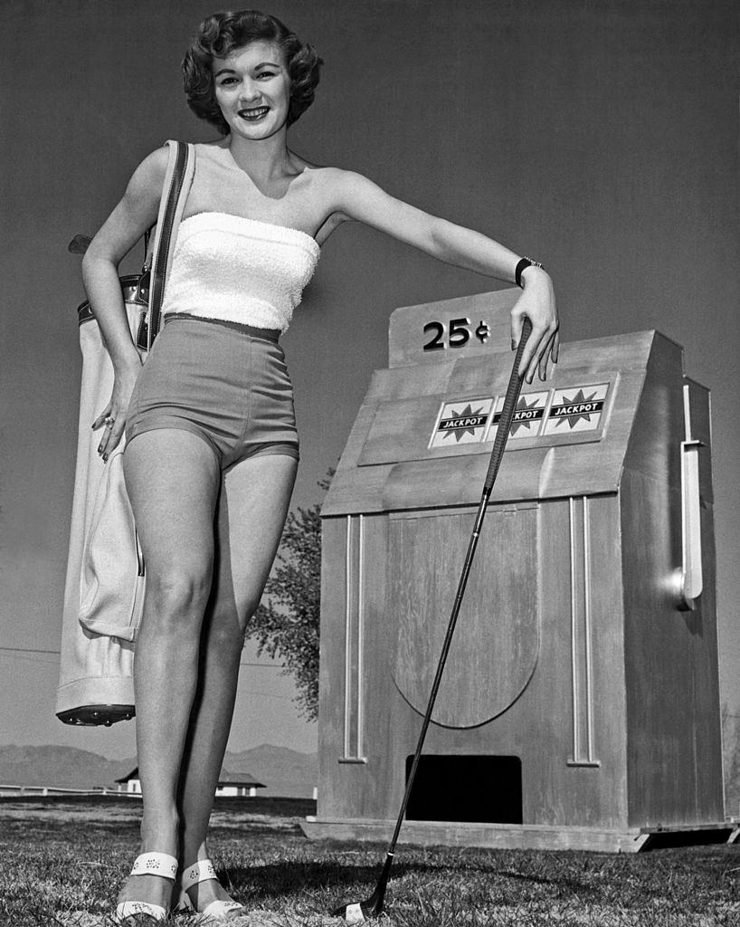 #54 Woman golfer in short shorts stands next to a model of a slot machine on the golf course, Las Vegas, 1956.