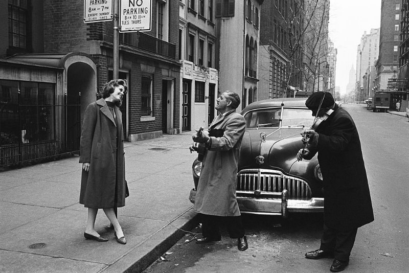 #16 Jane Fonda, leaning against a pole, smiling while listening to the street musicians playing for her.
