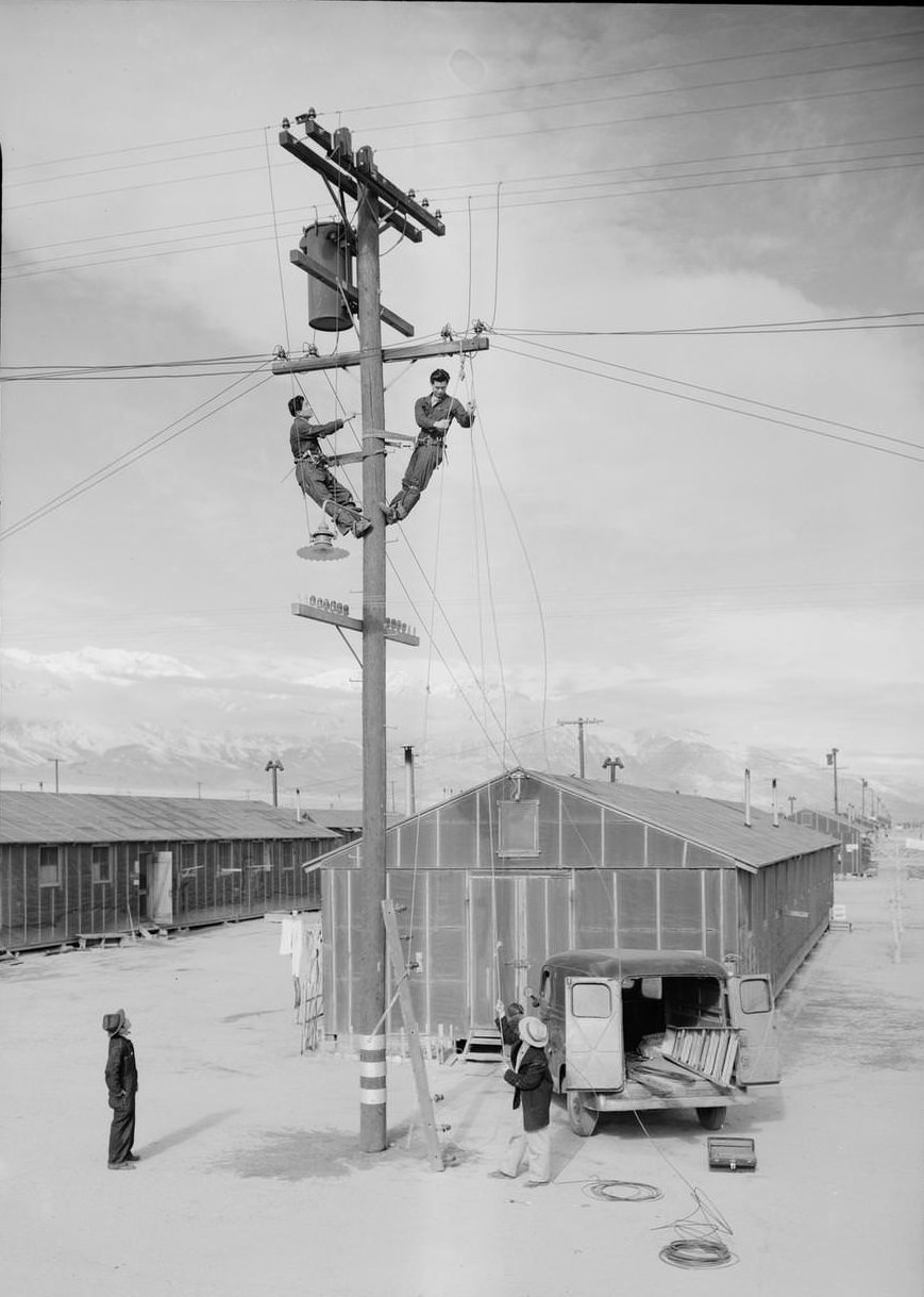 Electric line repair crew at work on electric pole next to building.